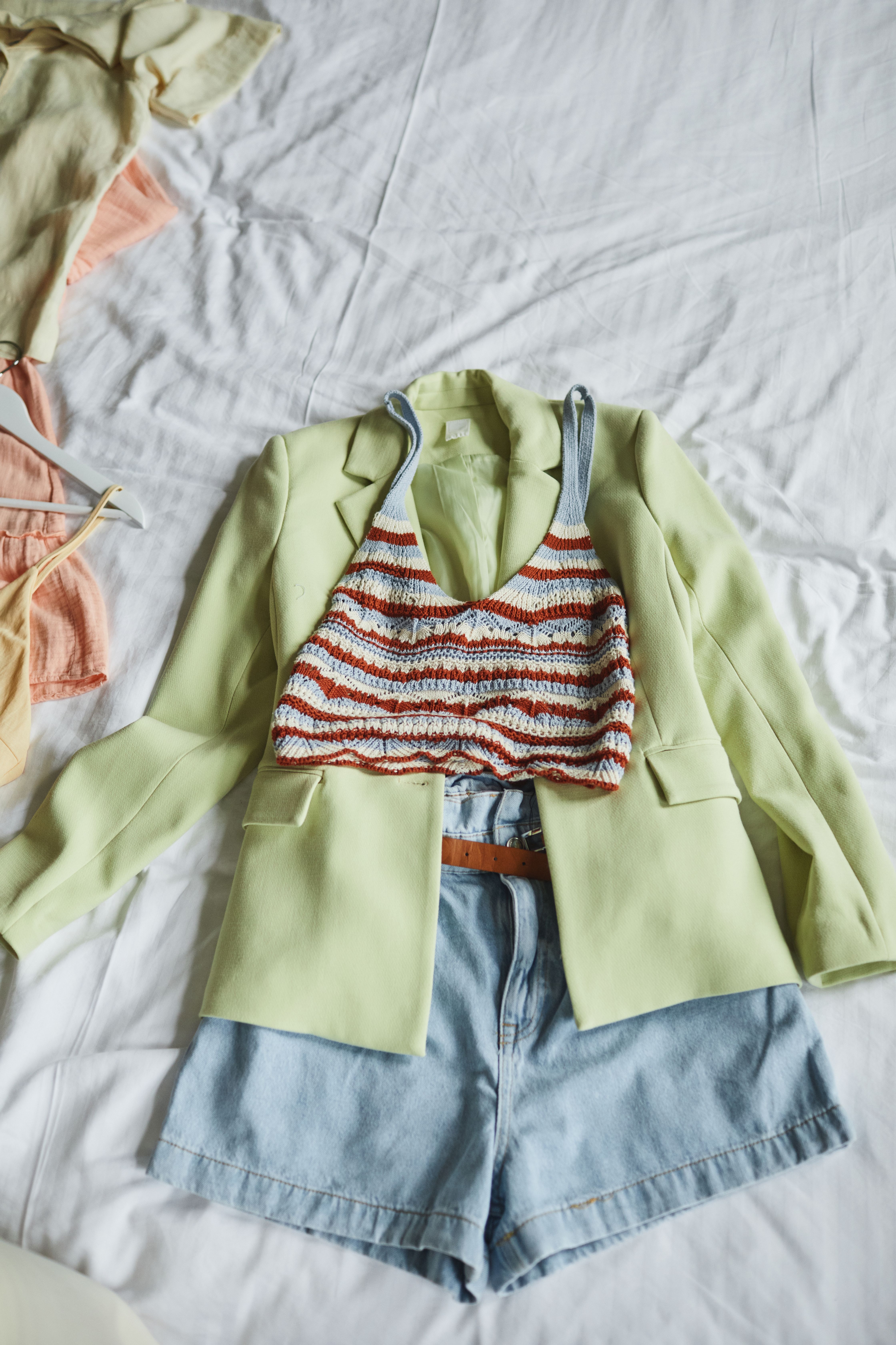 Pastel blazer, knitted top, and denim shorts arranged on a bed